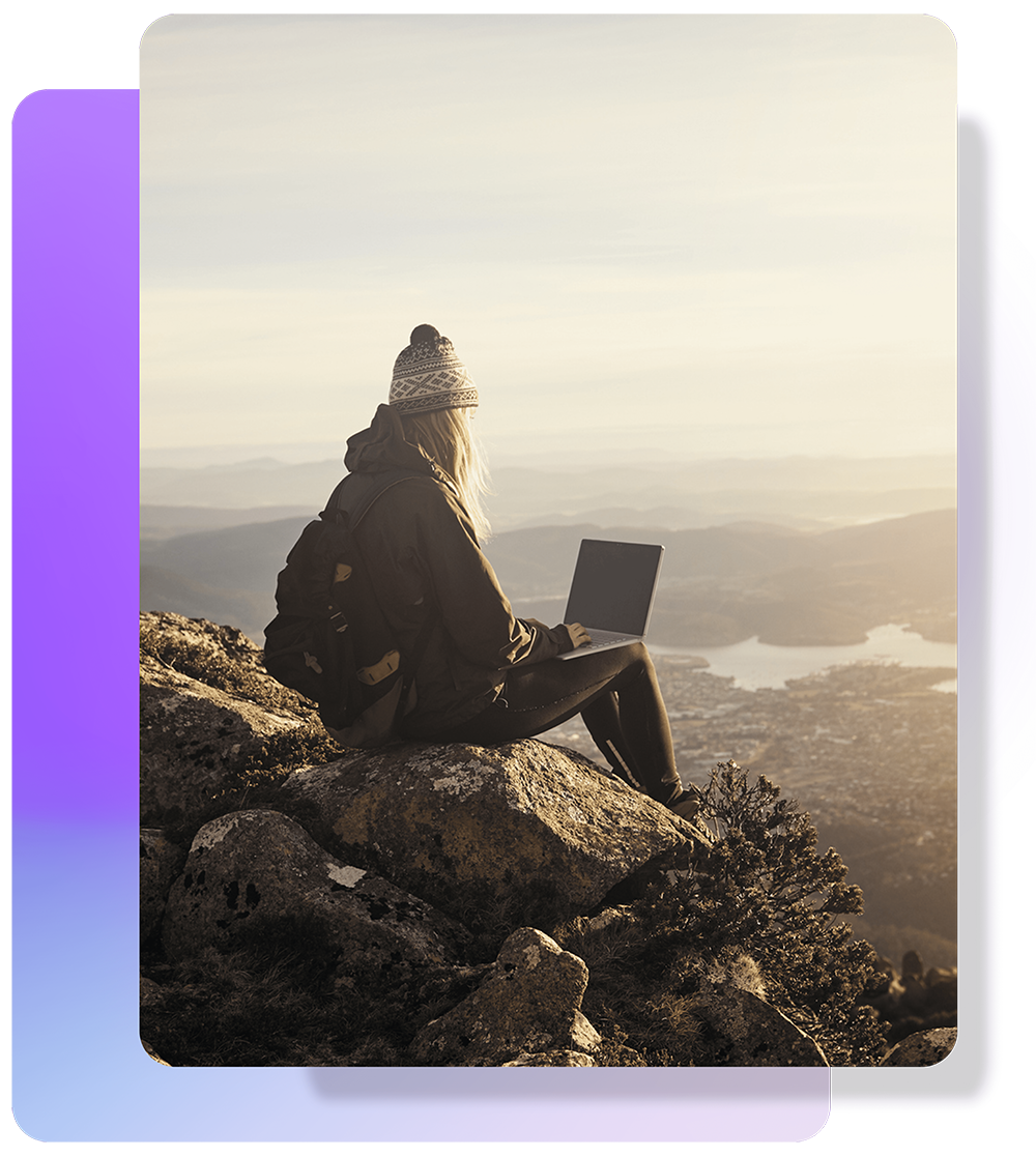 An image of a woman sitting on a mountain top and working on her computer.